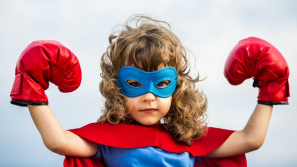 Un enfant est déguisé en superhéros. Il porte un masque bleu autour des yeux, une cape et des gants de boxe rouge. Ses poings sont levés de chaque côté de sa tête.