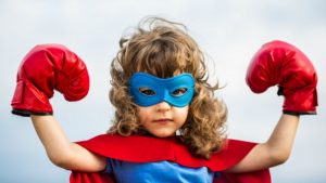 Un enfant est déguisé en superhéros. Il porte un masque bleu autour des yeux, une cape et des gants de boxe rouge. Ses poings sont levés de chaque côté de sa tête.