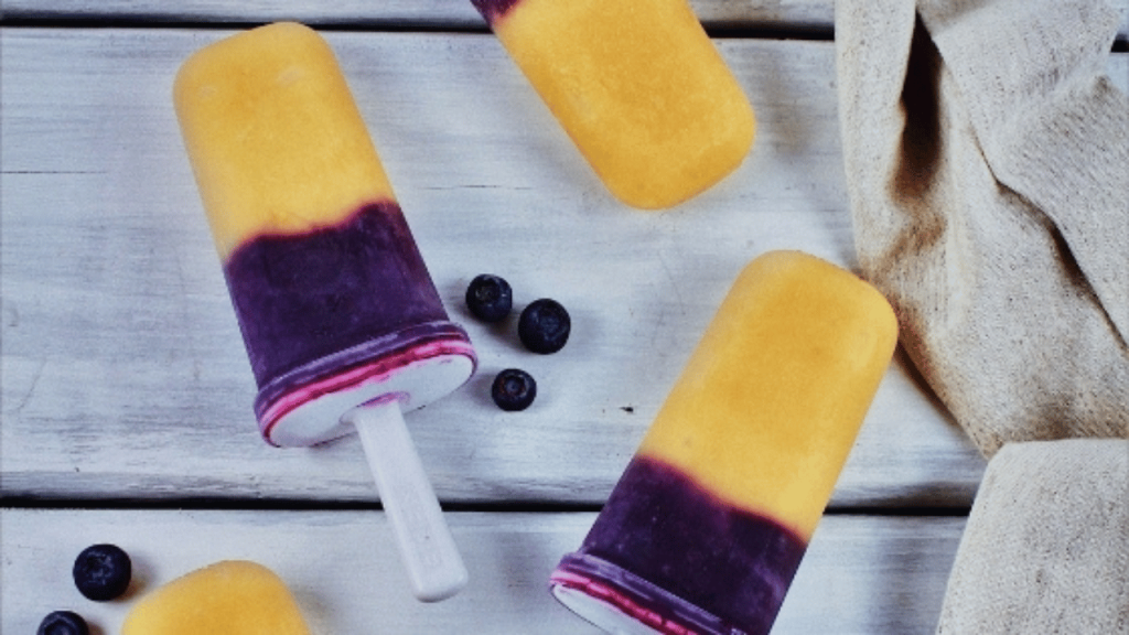 Popsicle maison 2 couleurs, mauve dans le bas et orange dans le haut sur une table en bois blanche et quelques bleuets frais