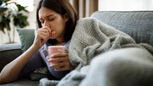 Femme assise sur un fauteuil portant son poing à son visage pour tousser en tenant une tasse de boisson chaude et enveloppée d'une couverture