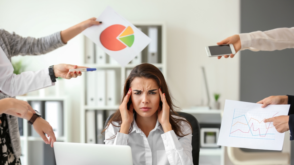 Une femme au travail se tenant la tête car elle est stressée devant un ordinateur et des collègues qui lui demande de faire des tâches