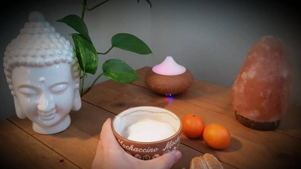 Table avec une ambiance zen, boisson chaude tenue par une main, un diffuseur d'huiles essentielles, une tête de Bouddha et une lampe de sel