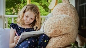 Petite fille assise sur un sofa qui écrit dans un cahier à côté d'un gros ourson en peluche