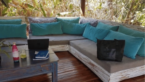 Sac de voyage noir posé sur un sofa extérieur adossé à la végétation tropicale et une table avec carnet de notes, ordinateur et bouteille d'eau.