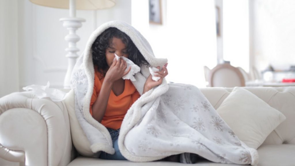 Une femme est assise sur un divan. Elle se mouche et a une couveture autour d'elle.