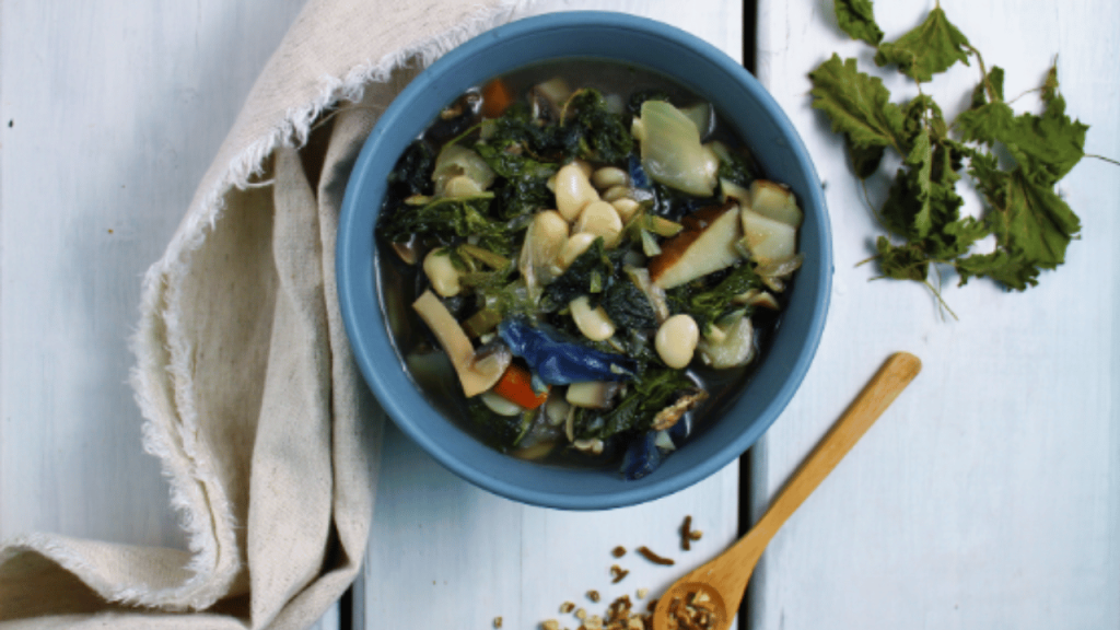 Soupe consistante avec bok choy et haricots blancs dans un bol bleu, une cuiller de bois posée sur la table blanche et des feuilles d'ortie en décoration.