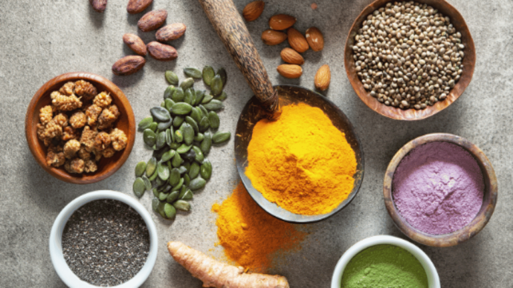 Des bols de poudre de superaliments de différentes couleurs et des bols de graines déposés sur une table avec des amandes et du curcuma frais